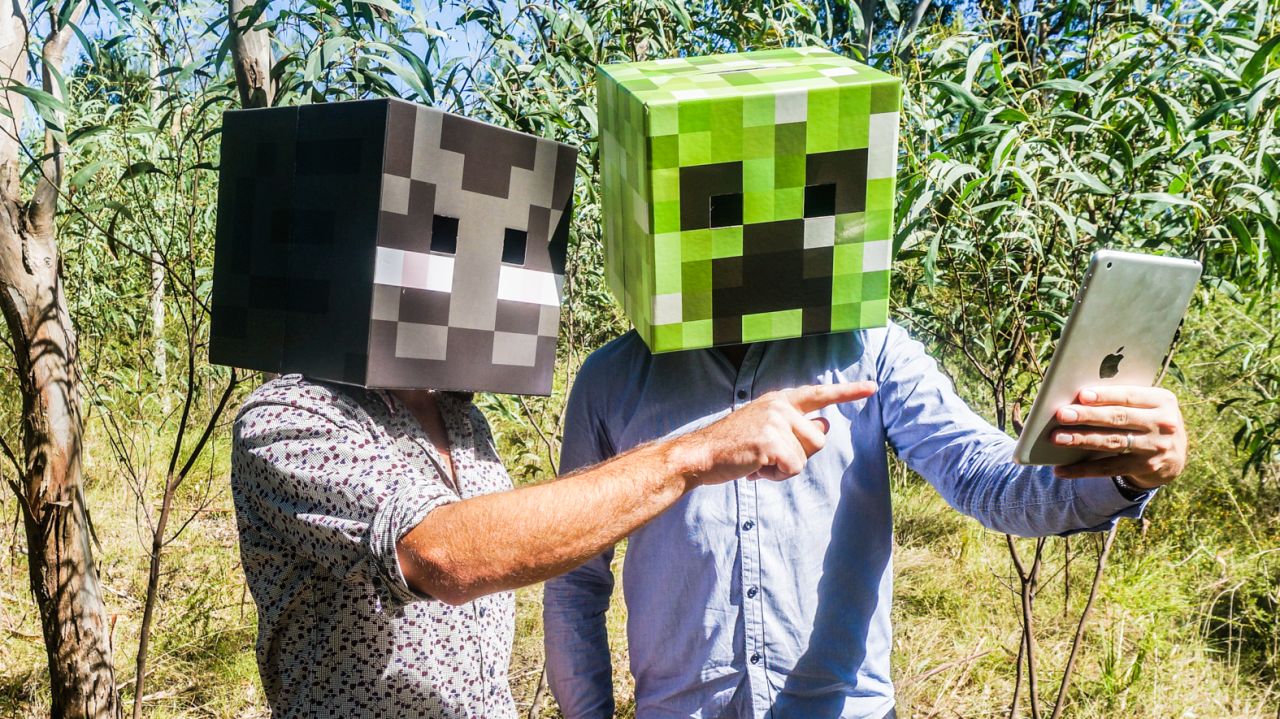 Two people wearing cardboard Minecraft heads in the bush, one pointing at a tablet