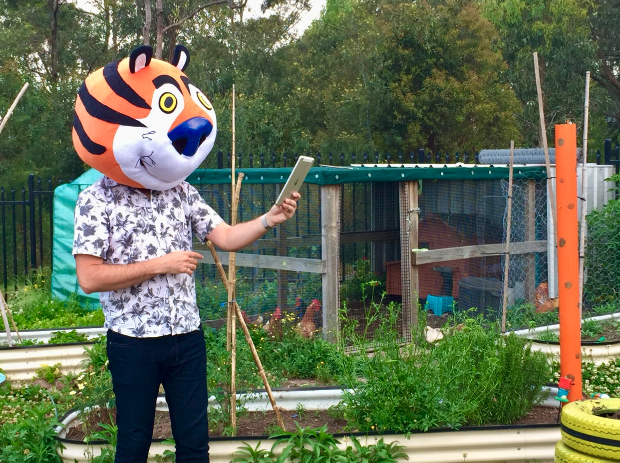 Craig in a tiger mask holding a tablet in a garden, gesturing toward a chicken coop