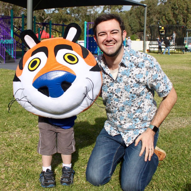 Craig kneeling beside a child wearing a large Frosties tiger mascot head in a playground